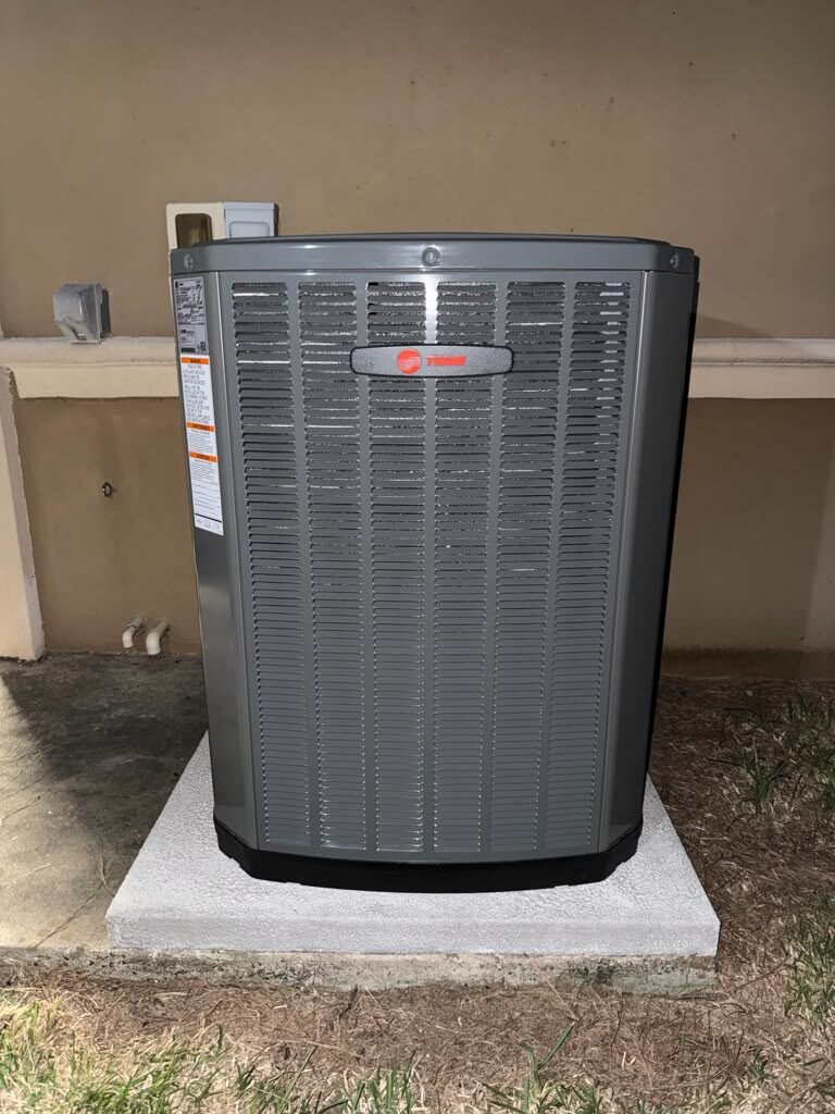 A Trane outdoor condensing unit on a new concrete pad beside a house wall.