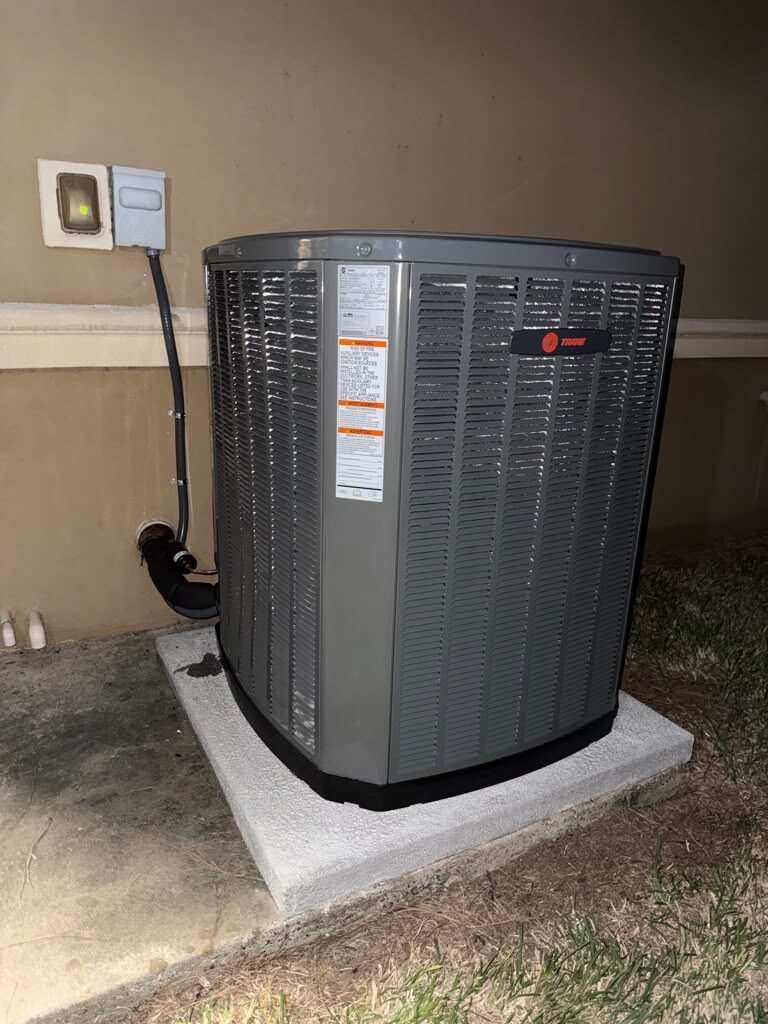 A Trane condensing unit on its pad at night, with the line set and electrical disconnect beside it.