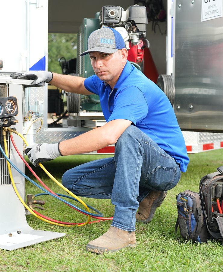 A technician crouched at a condensing unit with gauges and a tool bag, with the RF trailer behind.