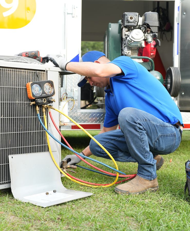 A technician bent over a condensing unit on the grass, adjusting gauge hoses.
