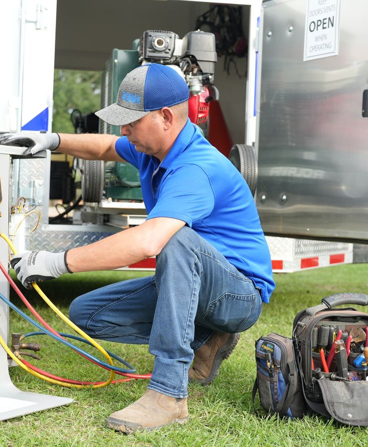 A technician crouched beside an outdoor condensing unit with gauge hoses connected, next to the RF service trailer.