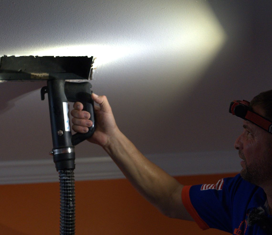 A technician with a headlamp vacuuming dust from a ceiling air register.